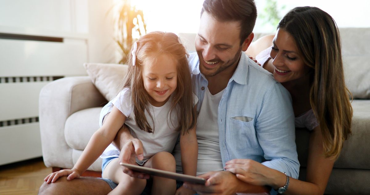 A father, mother, and young daughter smiling while watching videos on a tablet on the living room floor